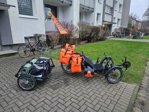 Drei Fahrräder mit Anhängern stehen auf einem gepflasterten Platz vor einem Wohngebäude.